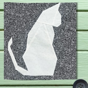 May include: A white origami cat design on a black and white patterned background. The cat is sitting, with its tail curved. The background has a repeating pattern of white squiggles. The image is on a green wooden surface.