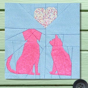 Könnte beinhalten: Ein Patchwork-Quadrat mit einem rosa Hund und einer Katze, die sich unter einem Herzen gegenübersitzen. Der Hund und die Katze bestehen aus rosa gemustertem Stoff, der vor einem hellblauen Hintergrund platziert ist. Ein Herz aus mehrfarbigem Stoff befindet sich über den Tieren.