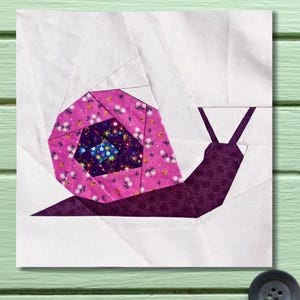 May include: A fabric art piece featuring a geometric snail design. The snail's shell is a vibrant pink with a floral pattern, and the body is a deep purple. The background is white, creating a striking contrast. The artwork is likely a quilt block.