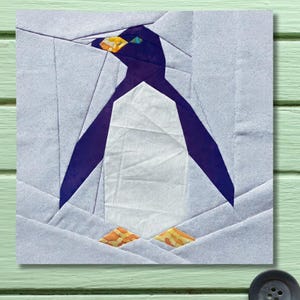 Peut inclure: Une œuvre d'art textile représentant un pingouin géométrique. Le pingouin a une poitrine blanche, un corps violet foncé et des pieds orange et jaunes. Le fond est bleu clair, et l'œuvre est sur un carré de tissu.