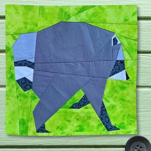 May include: A fabric quilt block featuring a stylized raccoon shape constructed from geometric pieces of blue and dark patterned fabric, set against a bright green background with a mottled texture and visible seam lines throughout the design.