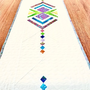 May include: A white table runner with a geometric design in shades of blue, green, purple, and orange. The design features a series of diamonds and squares arranged in a symmetrical pattern.