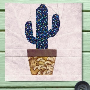 May include: A fabric art piece featuring a cactus in a pot. The cactus is a vibrant blue with a floral pattern, and the pot has a brown and gold design. The background is a light pink, and the artwork is mounted on a light green surface.