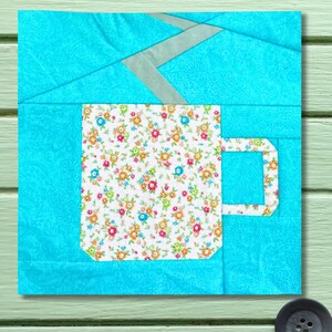 May include: A quilted fabric art piece featuring a coffee mug design. The mug is filled with a floral pattern of orange, pink, and green flowers on a white background. The background is a light blue color.