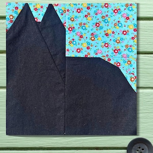 May include: A square fabric block with a patchwork design. The top section features a light blue floral pattern with red, pink, and yellow flowers. The bottom section is a dark navy blue fabric, with a geometric shape.