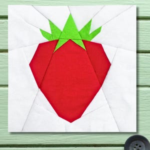 Peut inclure: Un bloc de tissu carré avec un motif géométrique de fraise rouge et de feuilles vertes. La fraise est centrée sur un fond blanc, le motif étant créé à partir de morceaux de tissu. Le bloc est probablement destiné au quilting ou à l'artisanat.