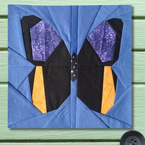 May include: A square, fabric patchwork design featuring a butterfly. The butterfly has black wings with orange and white accents, purple floral patterned sections, and a blue background. The butterfly's body is a black and white pattern.
