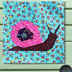 May include: A fabric block with a snail design. The snail's shell is pink and purple, and the body is brown. The background is a floral pattern in blue, red, and yellow. This handmade item is suitable for quilting and patchwork projects.