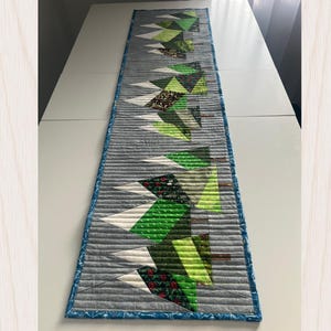 Peut inclure: Un chemin de table matelassé rectangulaire avec un fond gris et des coutures horizontales. Le motif présente une série d'arbres à feuilles persistantes stylisés dans des tons de vert, marron et blanc, avec une bordure à motifs bleus.