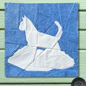 May include: A fabric square featuring a white cat silhouette on a blue background with a floral pattern. The cat is sitting on a white cloud-like shape. The design is created using patchwork techniques, showcasing a handmade, artistic style.