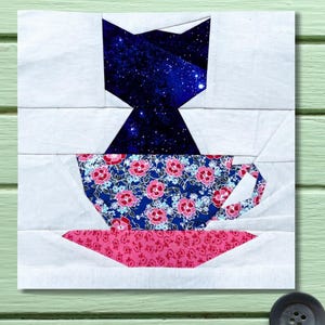 May include: A fabric block depicting a cat silhouette filled with a starry night sky. Below the cat is a teacup with a blue floral pattern and a pink saucer. The background is white.