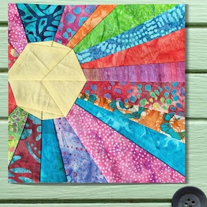 May include: A vibrant, handmade fabric artwork with a sunburst pattern. The sun's centre is a light yellow hexagon, with colourful rays in pink, blue, green, orange, and red. The fabric features diverse patterns, including polka dots and batik-style designs.