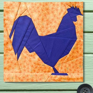 May include: A fabric square with a blue origami-style rooster on a floral orange background. The rooster is crafted from folded fabric, with detailed wings, tail, and comb. The background features a repeating pattern of small orange flowers.