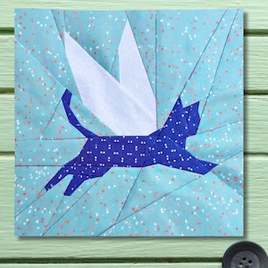 May include: A fabric art piece featuring a blue cat with white polka dots and white wings, set against a light blue background with red speckles. The cat is in mid-leap, giving the impression of flight. The artwork is a square quilt block.