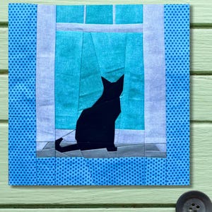 Può includere: Un'opera d'arte in tessuto raffigurante la silhouette di un gatto nero seduto in una finestra. La finestra ha tende turchesi e un davanzale grigio. L'opera è incorniciata con tessuto a pois blu e tessuto azzurro.