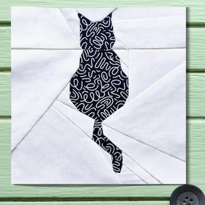 Pode incluir: Uma peça de arte têxtil branca e azul marinho com a silhueta de um gato. O gato é preenchido com um padrão azul marinho de redemoinhos brancos. O fundo é branco, com linhas geométricas criando um efeito de patchwork. A obra está sobre uma superfície verde clara.