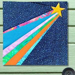 Könnte beinhalten: Ein quadratisches Stoffkunstwerk mit einem gelben Stern mit bunten diagonalen Streifen auf einem dunkelblauen Hintergrund. Die Streifen umfassen Rosa, Blau, Lila, Grün und Orange. Der Hintergrund hat ein gepunktetes Muster.
