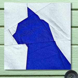 Puede incluir: Un bloque de tela cuadrado con un diseño geométrico. El diseño presenta una gran forma azul rodeada de piezas de tela blanca. La tela está doblada y cosida, creando un efecto de patchwork. El bloque está colocado sobre una superficie verde claro.