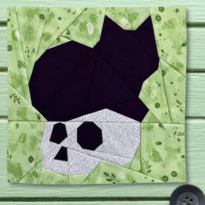 Könnte beinhalten: Ein quadratischer Stoff-Quiltblock mit einem Papier-Patchwork-Design, das eine einfarbige schwarze Katze hinter einem weiß-grau gemusterten Totenkopf zeigt. Der umgebende Rand besteht aus hellgrünem Stoff mit einem dezenten botanischen Druck aus Blättern und Sternen.