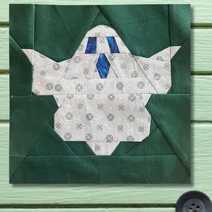 May include: A square fabric block featuring a geometric design of a teapot. The teapot is primarily white with a gray floral pattern and blue accents. The background is dark green, creating a striking contrast.