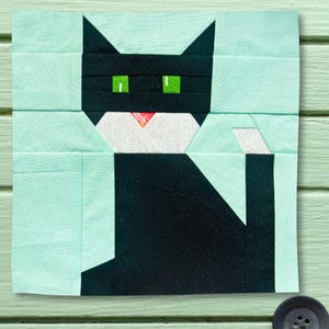 May include: A square fabric quilt block featuring a stylized black cat with bright green eyes, a white patterned muzzle, and a small pink nose. The cat is set against a solid light mint green background, constructed from multiple rectangular fabric pieces.