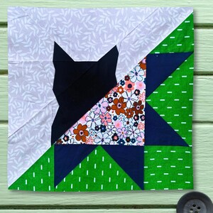 Könnte beinhalten: Ein quadratischer Stoff-Quiltblock mit einem zusammengesetzten Design. Das Muster enthält eine einfarbige schwarze Katzensilhouette, einen Abschnitt aus weißem Stoff mit hellgrauem Blattdruck, einen Blumendruck mit rosa und braunen Blüten sowie leuchtend grünen Stoff mit weißen Strichen.