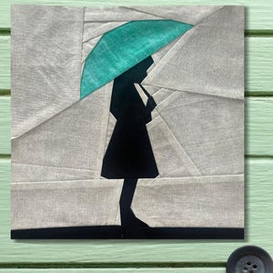 Puede incluir: Una obra de arte textil que representa la silueta de una persona con un paraguas. El paraguas es turquesa y la persona es negra. El fondo está compuesto por formas geométricas grises y blancas, creando un diseño abstracto moderno.