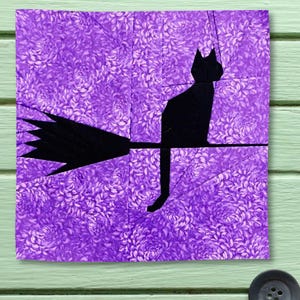 Könnte beinhalten: Ein quadratischer Stoff-Quiltblock mit der schwarzen Silhouette einer Katze, die auf einem Besenstiel sitzt. Der Hintergrund besteht aus violettem Stoff mit einem tonalen Blumenmuster. Das Design besteht aus mehreren Stoffteilen.