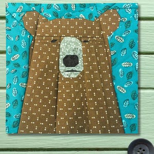 May include: A square fabric block featuring a bear face design. The bear is brown with white polka dots, a light green nose, and a black snout. The background is turquoise with white leaf patterns. The block is likely part of a quilt or other textile project.