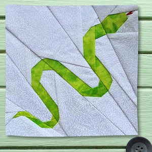 May include: A square fabric quilt block featuring a paper-pieced design of a winding green snake. The snake is constructed from bright green fabric with subtle yellow variations, set against a background of white fabric with a gray floral pattern.