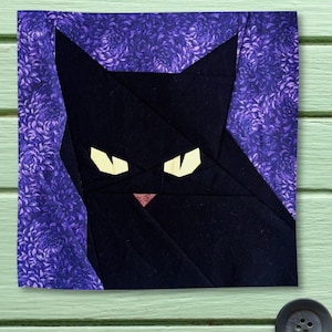 May include: A square fabric quilt block featuring a stylised black cat head constructed from geometric fabric pieces. The cat has pointed ears, pale yellow eyes, and a small pink nose, set against a background of deep purple floral patterned fabric.