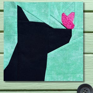 May include: A geometric quilt block featuring a black cat silhouette with a pink heart. The cat is set against a light green background. The design is a modern take on a traditional quilt pattern.