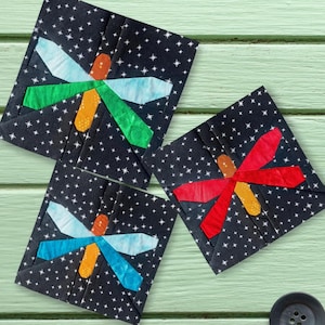 May include: Three square fabric coasters with dragonfly designs. Each coaster features a black background with white star patterns and a colorful dragonfly applique. The dragonflies have wings in shades of blue, green, and red.