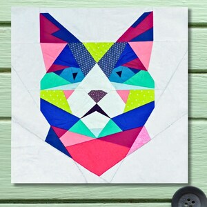Könnte beinhalten: Ein farbenfroher geometrischer Katzenkopf-Quiltblock. Das Design zeigt ein Patchwork aus Dreiecken in Rosa, Blau, Grün und Weiß. Das Gesicht der Katze ist auf einem weißen Hintergrund zentriert.