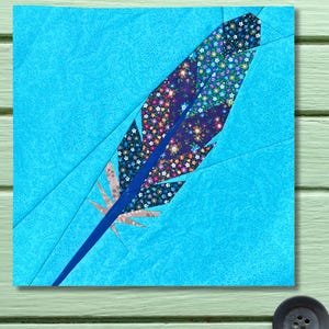 May include: A colourful, quilted feather design. The feather has a dark blue stem and is filled with floral fabric in shades of blue, purple, and orange. The background is a light blue fabric with a paisley pattern. The design is square and set against a light green surface.
