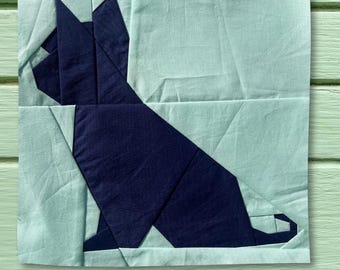 以下が含まれることがあります： 明るいティール色の背景に、ネイビーブルーの猫のシルエットが描かれた正方形の布ブロック。 猫は幾何学的な、折り紙のようなスタイルで描かれています。 ブロックは、パッチワークのデザインを示す、ステッチされた布のピースで作られています。