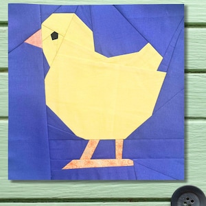 May include: A patchwork design of a yellow chick on a blue background. The chick has a pink beak, black eye, and patterned legs. The square design is likely a quilt block, with geometric shapes forming the chick.