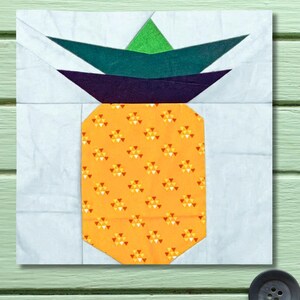 Puede incluir: Un bloque de tela de piña con un fondo azul claro. La piña tiene un cuerpo naranja con un patrón repetido de triángulos amarillos y naranjas. La parte superior presenta hojas verdes, verde oscuro y moradas.