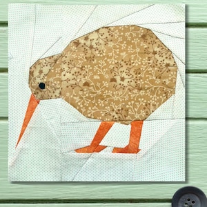 May include: A square fabric quilt block featuring a stylized kiwi bird. The bird's body is a patchwork of brown floral fabric, with orange beak and legs. The background is white with small blue dots.