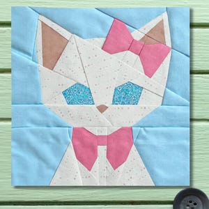Puede incluir: Un bloque de tela con un diseño de gato. El gato tiene una cara blanca con lazo rosa, orejas y pajarita. Los ojos son azules con un estampado floral. El fondo es azul claro.