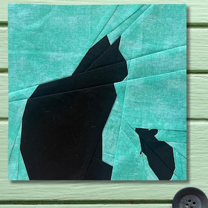 Könnte beinhalten: Ein quadratisches Stoffkunstwerk mit einer schwarzen Katzen-Silhouette vor einem türkisfarbenen Hintergrund. Die Katze besteht aus geometrischen Formen, und der Hintergrund hat ein strukturiertes Aussehen. Das Kunstwerk wird auf einer hellgrünen Oberfläche präsentiert.