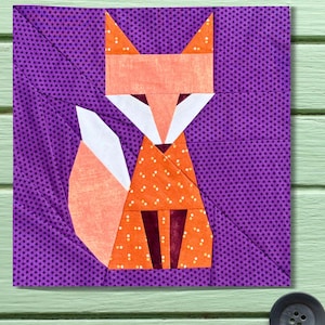 以下が含まれることがあります： 幾何学的なキツネのデザインが施された正方形の布ブロック。キツネは、オレンジ、白、茶色の布地で構成され、紫色の水玉模様の背景に配置されています。キツネの尾は白とピーチ色です。ブロックのサイズは約20cm四方です。