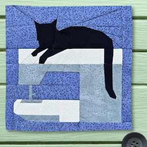 May include: A square fabric quilt block featuring a black cat silhouette resting on top of a sewing machine. The background is composed of blue fabric with a delicate white leaf pattern, while the sewing machine is pieced from white and light gray fabric.