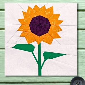 May include: A fabric square featuring a paper-pieced sunflower design. The sunflower has a purple center, orange petals with yellow accents, and a green stem and leaves. The background is white with subtle geometric lines.