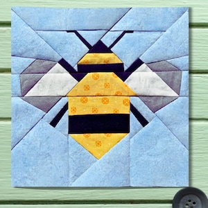 May include: A square quilt block featuring a geometric design of a bee. The bee has a yellow body with black stripes and a yellow head. The wings are white and gray, set against a light blue background. The quilt block is on a light green surface.