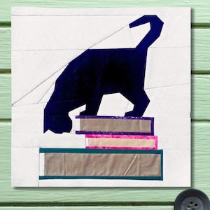 Könnte beinhalten: Ein geometrischer Quiltblock mit einer dunkelblauen Katzen-Silhouette auf einem Stapel von drei Büchern. Die Bücher haben violette, rosa und türkisfarbene Akzente. Der Hintergrund ist weiß mit geometrischen Linien.