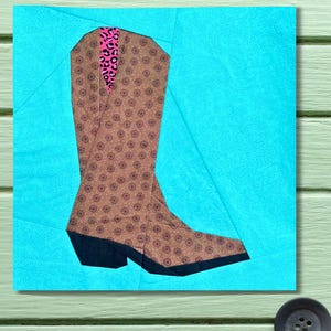 以下が含まれることがあります： ターコイズブルーの背景に、ピンクのヒョウ柄のアクセントが付いた茶色と黒のカウボーイブーツ。 ブーツは詳細な花柄と尖ったつま先が特徴です。 アートワークは正方形の幾何学模様です。