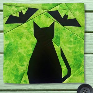 May include: A square fabric quilt block featuring a pieced design of a solid black cat sitting in the center with two black bats flying above it, all set against a vibrant, mottled lime green background with visible geometric seam lines.