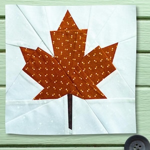 May include: A square fabric quilt block featuring a central maple leaf design. The leaf is constructed from brown fabric with a small white dot pattern, set against a background of light blue fabric with a subtle white speckled texture.