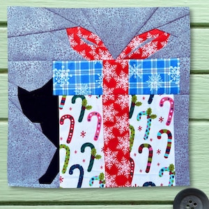 May include: A square fabric quilt block featuring a black cat silhouette next to a gift box. The gift is wrapped in white fabric with candy canes, a blue plaid ribbon with white snowflakes, and a red ribbon with white snowflake patterns.